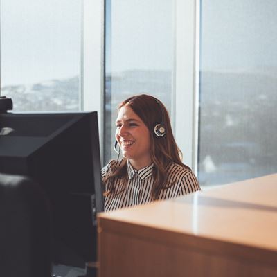 En kvinde som sidder i et kontor forand en computer med headset på. 