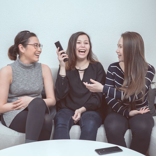 Tre teenagere sidder i en sofa med en fjernbetjening og telefon. 