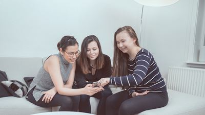 Tre teenagere sidder op en sofa og kigger i en mobiltelefon.