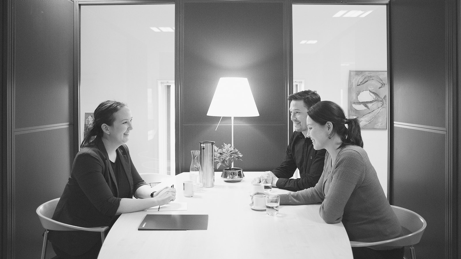 Et kundemøde set fra siden, hvor en rådgiver og to kunder sidder ved et bord. 